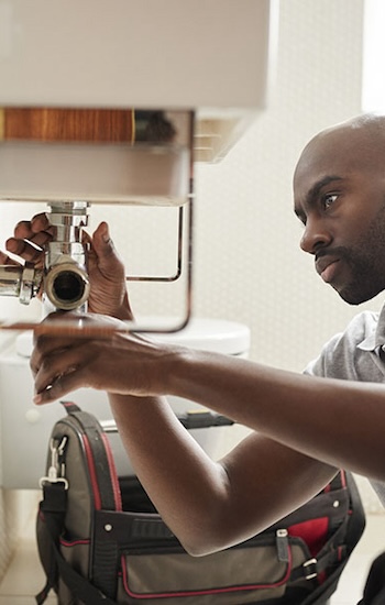 Young black male plumber sitting on the floor fixing a bathroom sink, seen from doorway Young black male plumber sitting on the floor fixing a bathroom sink, seen from doorway